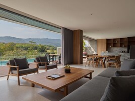 Una sala de estar moderna con una amplia mesa de centro de madera, sofá gris y comedor con sillas de madera. Un gran ventanal ofrece vistas a una piscina y montañas.
