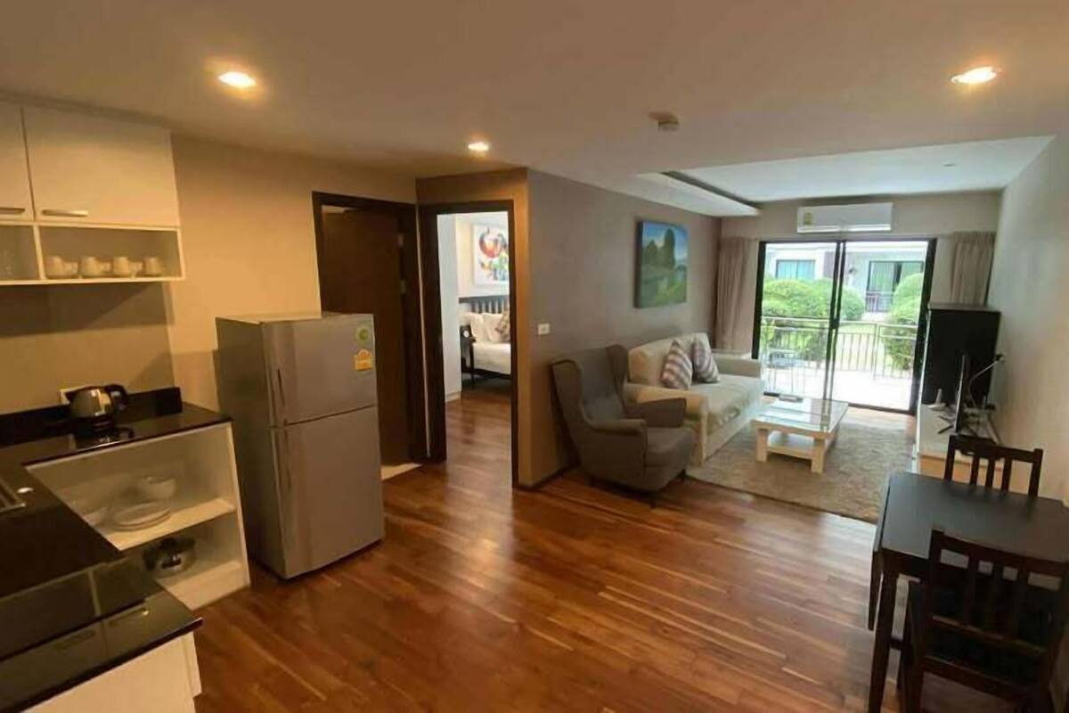 A modern kitchen with a refrigerator, stove, and cabinets. There is a dining area with a table and chairs. A living room with a sofa and a TV is visible through an open door.