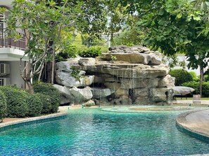 A pool with a waterfall feature surrounded by lush greenery and a multi-story building. - The Title Rawai Aparthotel by HSS (Rawai)