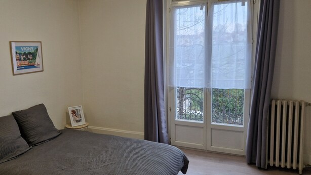 A bedroom with a bed, two framed pictures on the wall, a window with curtains, and a radiator. - 26 m2 studio with separate kitchen and balcony, opposite the station (Vichy)