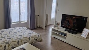 A hotel room with a bed, a TV on a stand, a radiator, and a window with curtains. - 26 m2 studio with separate kitchen and balcony, opposite the station (Vichy)