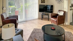 Ein Wohnzimmer mit Fernseher, zwei Ledersesseln, einem Couchtisch, einer Lampe und einer Schiebetür zum Garten.