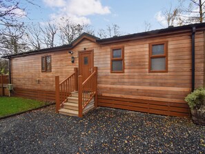 A wooden cabin with a wooden staircase leading to a door, a small porch, and a gravel driveway. - Attractive two bedroom, two bathroom lodge, with hot tub. (Water Yeat, Cumbria)
