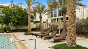 Zona junto a la piscina con sillones de descanso, palmeras y un edificio con amplias ventanas.