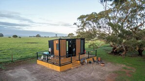 Una cabaña moderna y compacta con balcón, ubicada en un campo cubierto de césped con árboles al fondo.