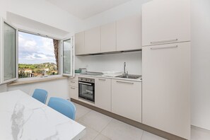 A modern kitchen with white cabinets, a stove, oven, and a sink. There is a dining table with chairs and a window with a view of buildings and trees. - Il Cavo Suite 1 - un nido Vista Mare (Rio Marina)