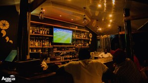 A bar area with a television displaying a sports game, shelves stocked with bottles, and a bar counter. - Acacia Boutique Hotel (Mbarara)