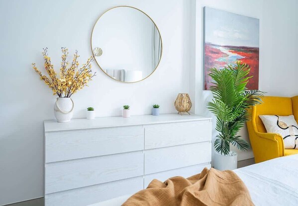A bedroom with a white dresser, a round mirror, a plant, a yellow chair, and a painting on the wall.