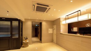 A modern hotel reception area with a polished floor, a reception desk, a wall-mounted air conditioner, and a hallway leading to other rooms. - AVENUE S HOTEL (Siheung)