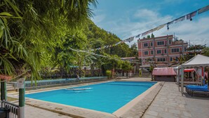 En swimmingpool med en stenbelagd poolkant, omgiven av frodig grönska och en byggnad i bakgrunden.