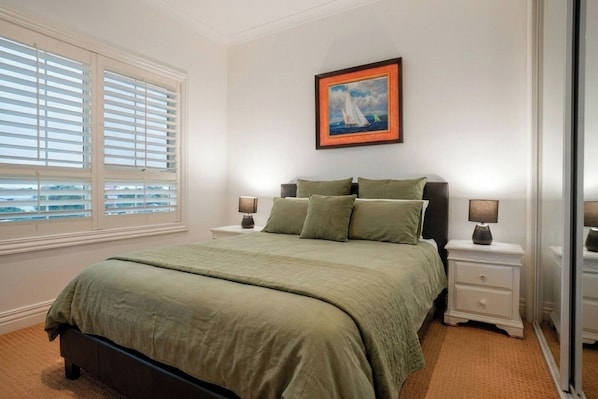 A bedroom with a bed, two bedside tables, a nightstand, a window with blinds, and a framed picture on the wall.