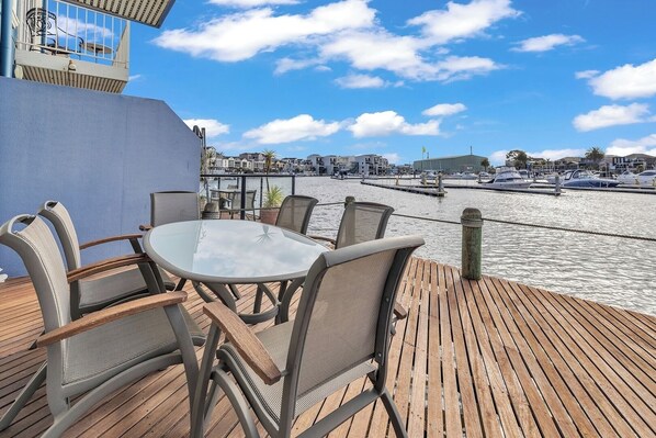 Una terraza de madera con una mesa redonda y sillas, con vistas a un puerto deportivo con barcos y edificios al fondo.