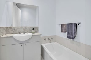 Un baño con lavamanos blanco, dos grifos, un espejo, una botella de champú, una bañera y dos toallas colgadas en un perchero.
