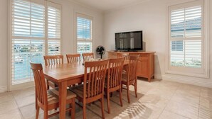 A dining area with a wooden table and chairs, a television on a wooden cabinet, and large windows with blinds.