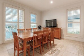 A dining area with a wooden table and chairs, a television on a wooden cabinet, and large windows with blinds.