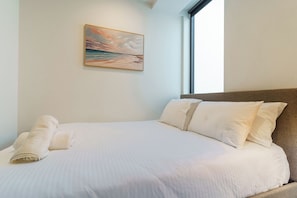 Ein ordentlich bezogenes Bett mit weißen Leinen und Kissen, ein aufgerolltes Handtuch und ein Strandbild an der Wand.