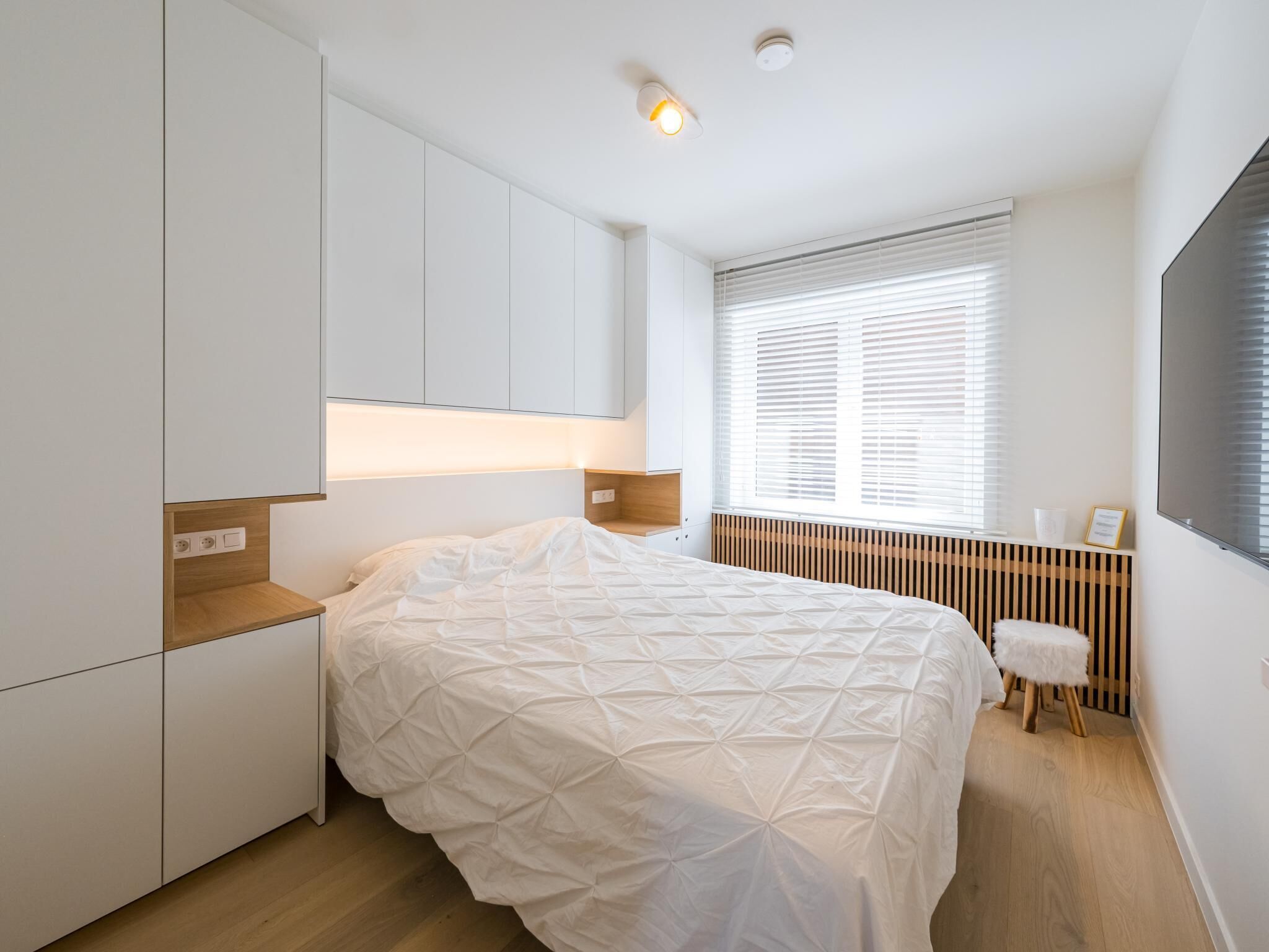 A modern bedroom with a bed, built-in wardrobe, bedside table, and a window with blinds.