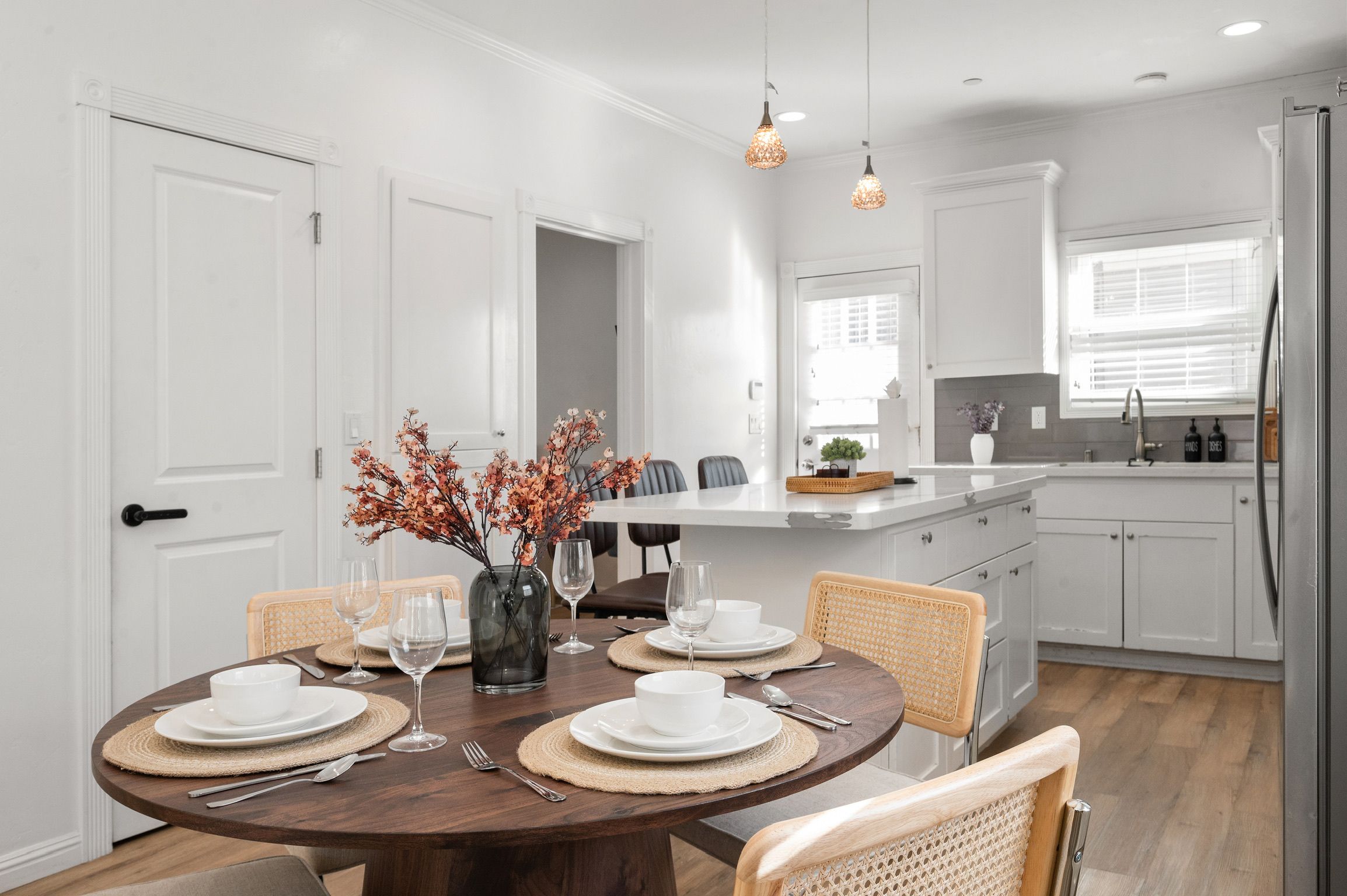 A dining area with a round table set for four, surrounded by wicker chairs, a vase with flowers, and pendant lighting.