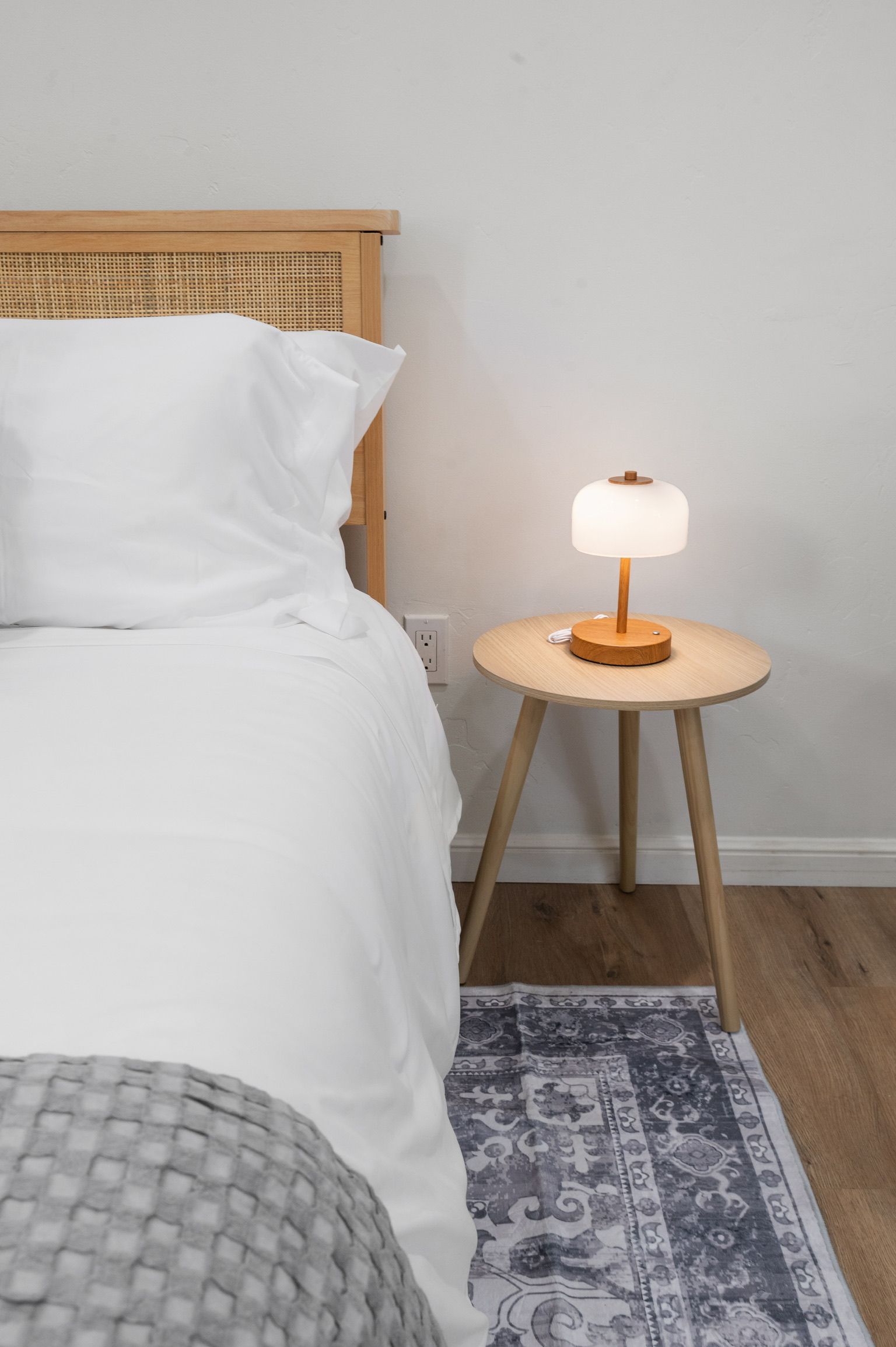 Ein Bett mit weißer Bettwäsche, ein runder Nachttisch mit Lampe und ein gemustertes Teppich auf Holzdielen.