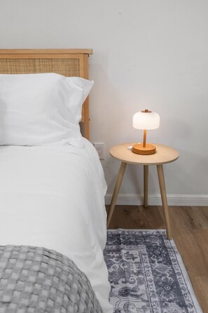 Ein Bett mit weißer Bettwäsche, ein runder Nachttisch mit Lampe und ein gemustertes Teppich auf Holzdielen.