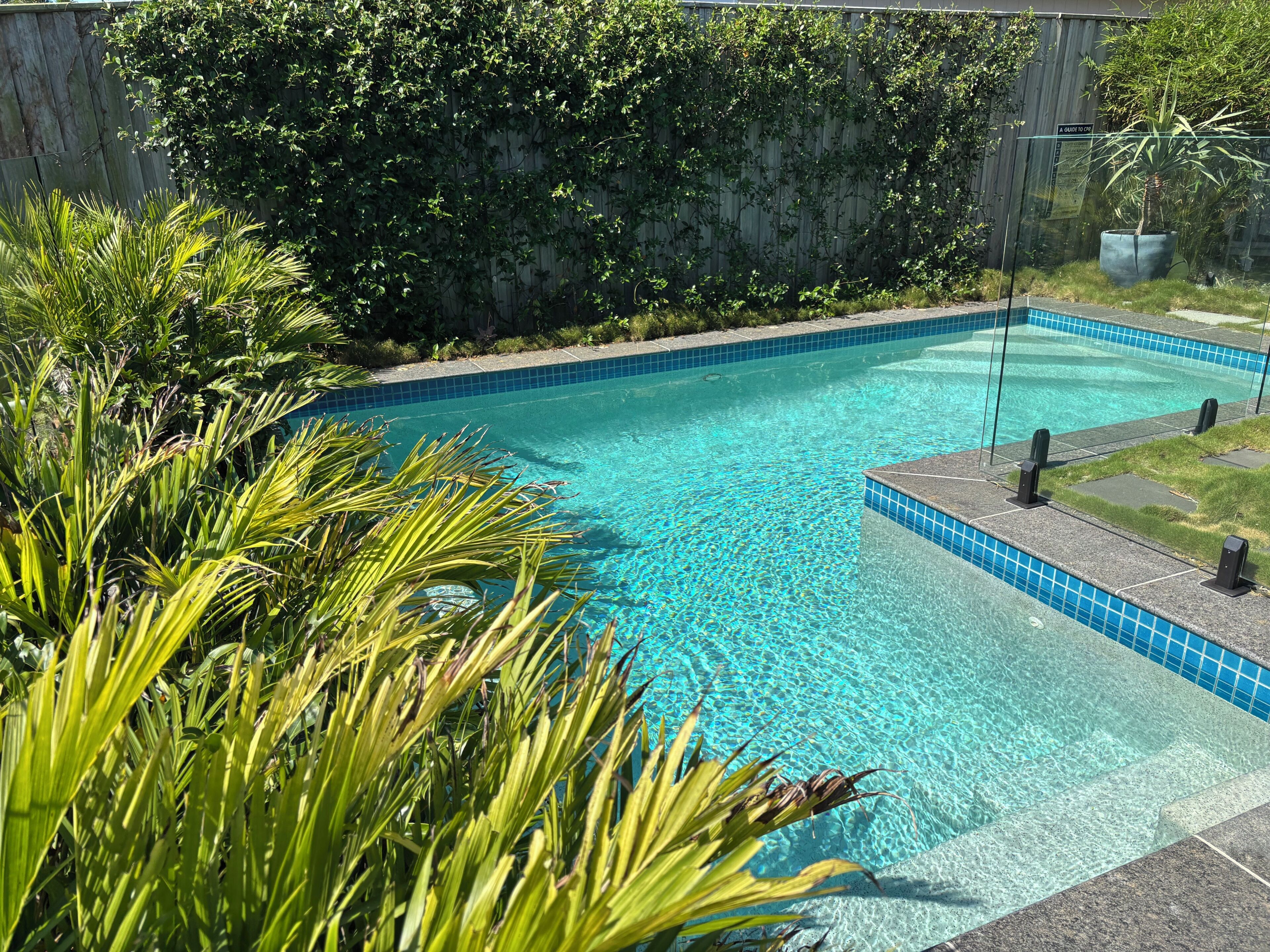 A clear blue swimming pool with a tiled edge, surrounded by lush greenery and a wooden fence.