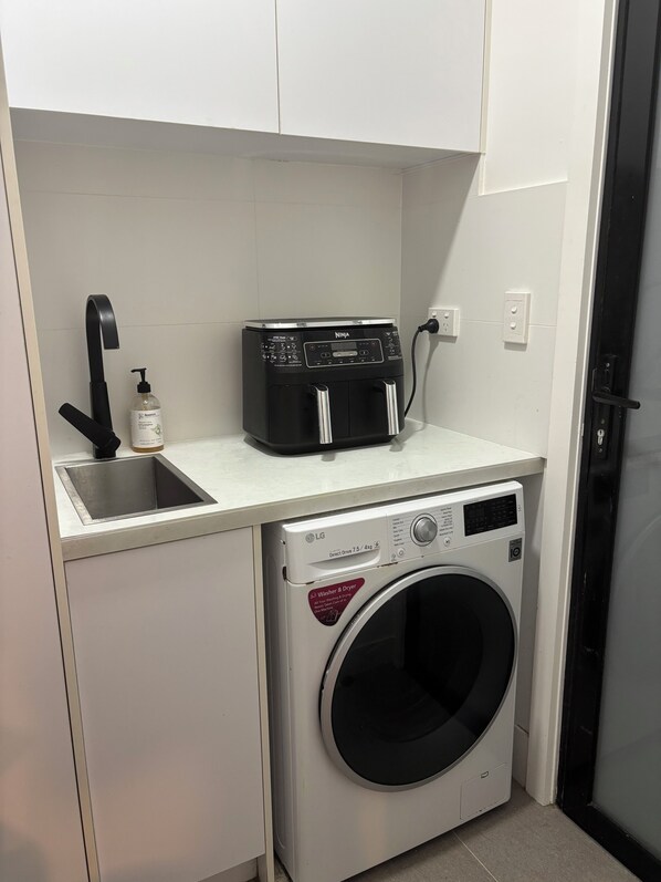 Uma área de lavanderia compacta, com máquina de lavar, pia e um eletrodoméstico preto na bancada.