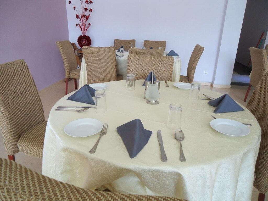 Un espace repas avec des tables rondes disposées pour accueillir quatre personnes, chacune étant recouverte d’une nappe blanche et agrémentée de chaises en osier marron, ainsi que de la vaisselle de base, comprenant des assiettes, des couverts et des serviettes.