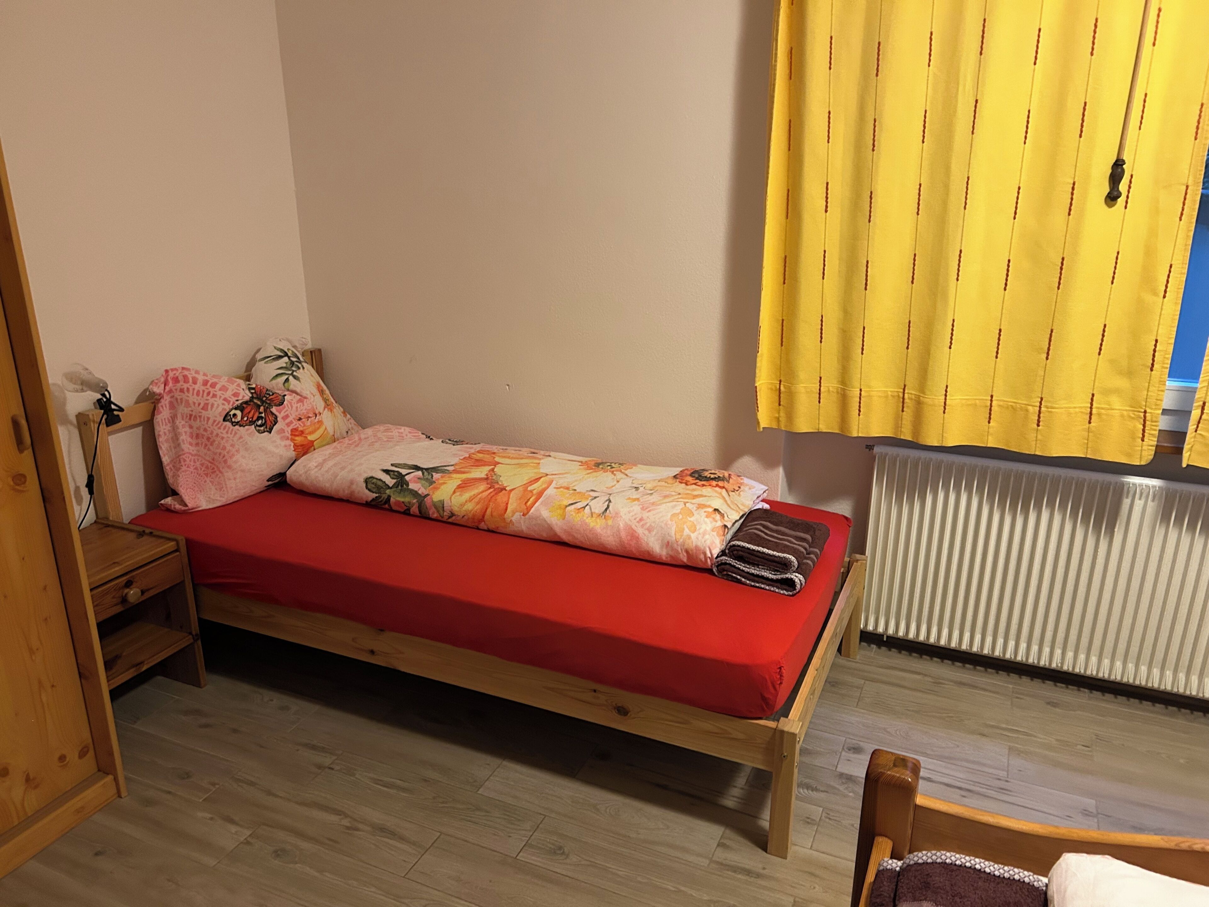 A small, single-bed room with a red mattress, wooden headboard, and a small wooden nightstand.