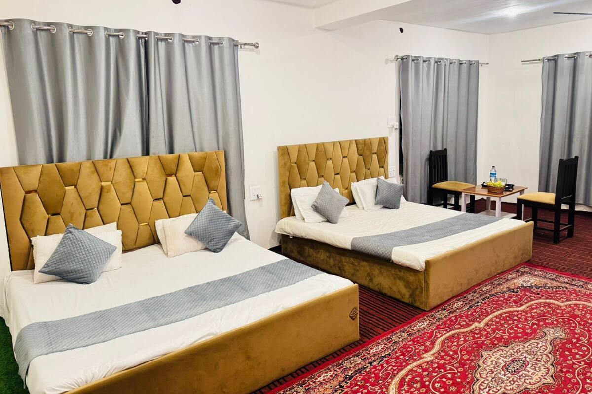 A hotel room with two beds, grey curtains, a wooden headboard, a red patterned carpet, and a small table with a bottle and fruits.