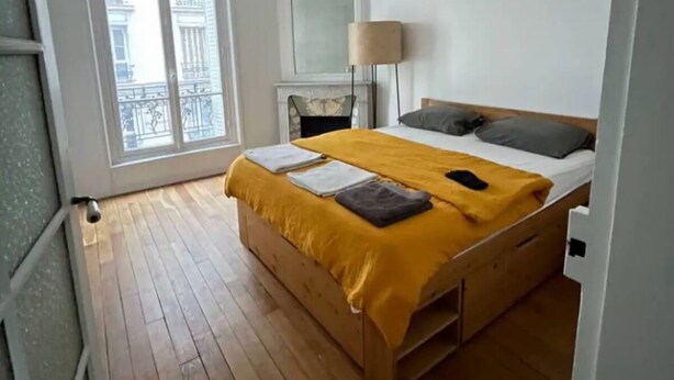 A bedroom with a wooden floor, a bed with a yellow blanket, a bedside lamp, and a view of buildings through a window. - Cosy Paris apartment 5 minutes from Bastille! Ideal for sightseeing! (Paris)