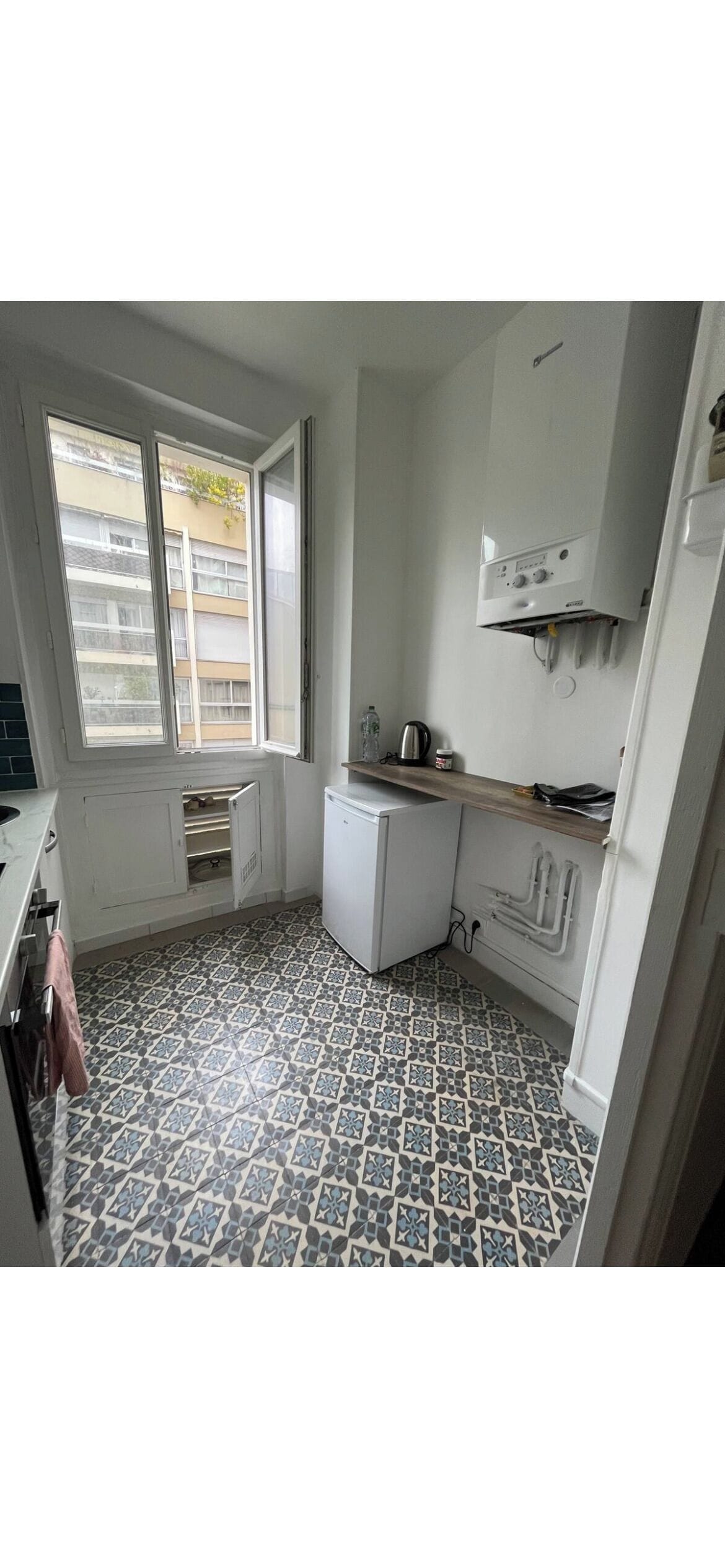 A compact kitchen with a window, white cabinets, a small refrigerator, and patterned floor tiles.