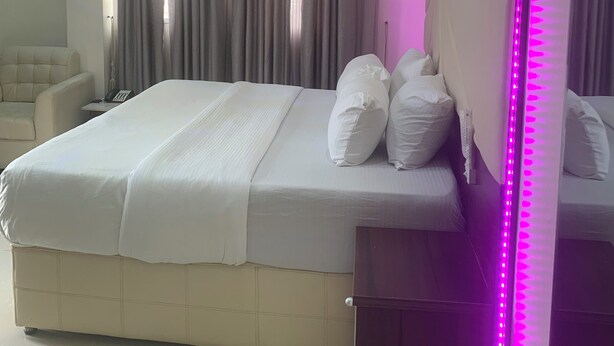 A neatly made bed with white bedding and pillows, a bedside table with a phone, and a large mirror with pink LED lights. - City Residence Lagos (Lagos)