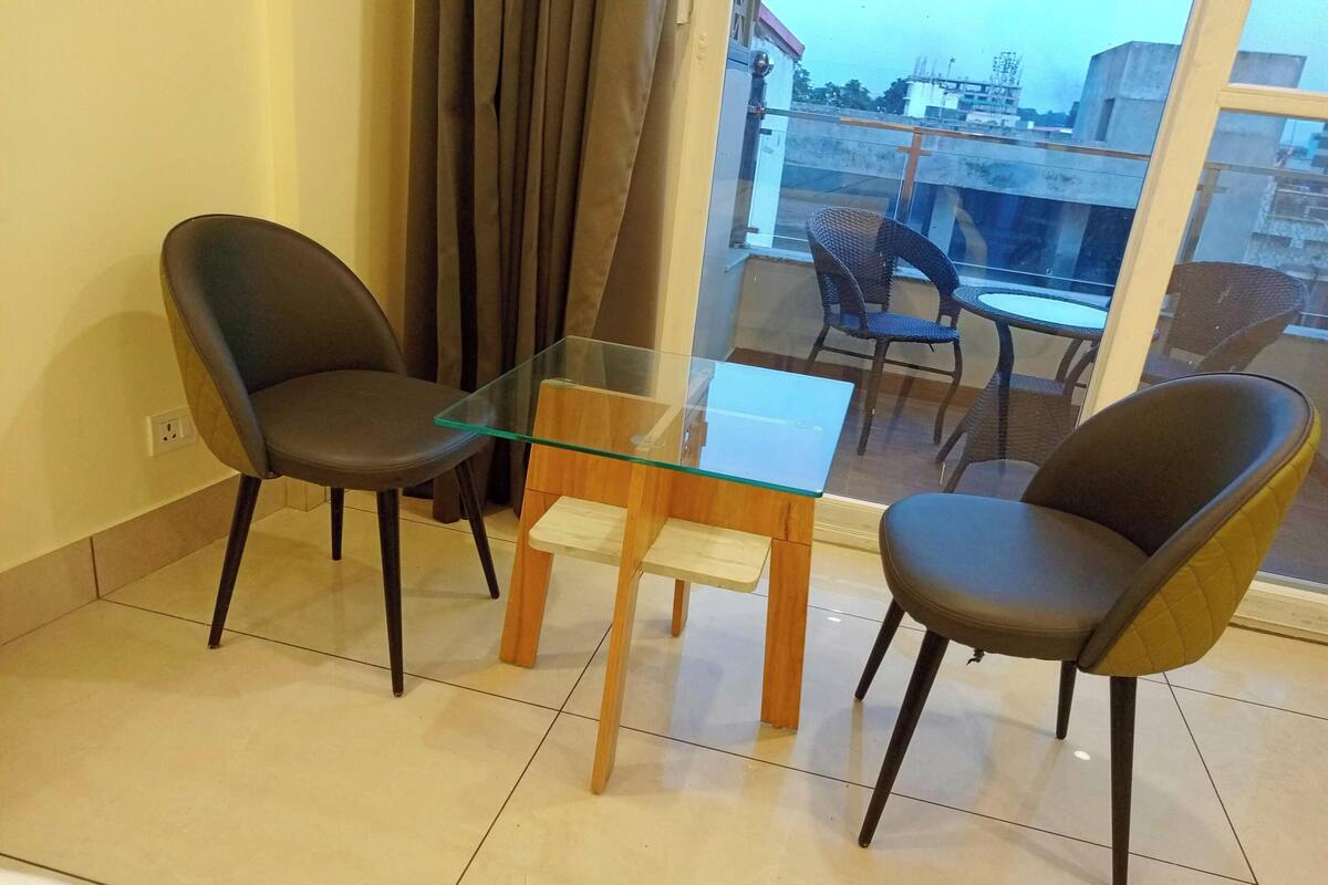 Un balcón con una mesa de vidrio y dos sillas, vistas de la ciudad y una ventana con cortinas.