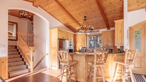 A kitchen with wooden cabinets, a central island with bar stools, a chandelier, and a stainless steel refrigerator. - New! Bearadise ~ The Retreat (Big Bear Lake)