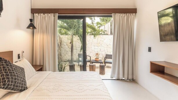 A bedroom with a bed, a nightstand, a lamp, a wall-mounted shelf, and a sliding glass door leading to an outdoor area with a table and chairs. - 2br Jungle Retreat with Private Pool (Tulumn)