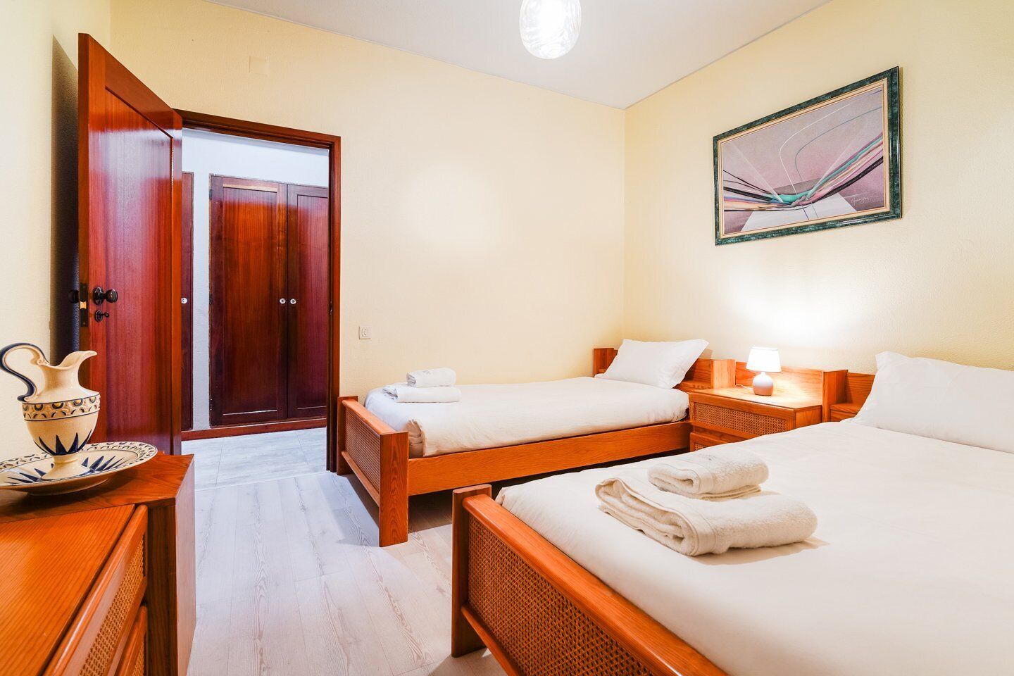 Een hotelkamer met twee houten bedden, een nachtkastje van hout, een bijzettafeltje, een abstract schilderij aan de muur en een deur met houten frame.