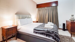 Een net opgemaakt bed met witte lakens en een donker, getextureerd dekbed. Er staat een houten nachtkastje met een lamp en een klein lade. Een houten dressoir met een decoratieve vaas bevindt zich rechts.