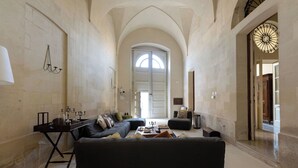 Amplia sala de estar con un sofá negro grande, una mesa de centro y una chimenea. Existe un arco grande que da a otra habitación, y un candelabro colgando del techo.