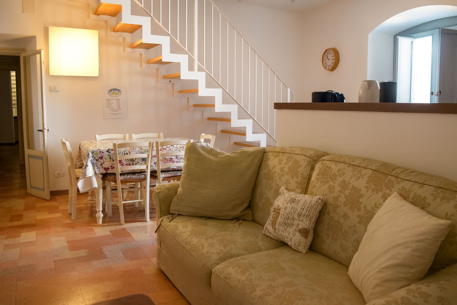 A living room with a beige sofa, a dining table with chairs, and a staircase with white railings.