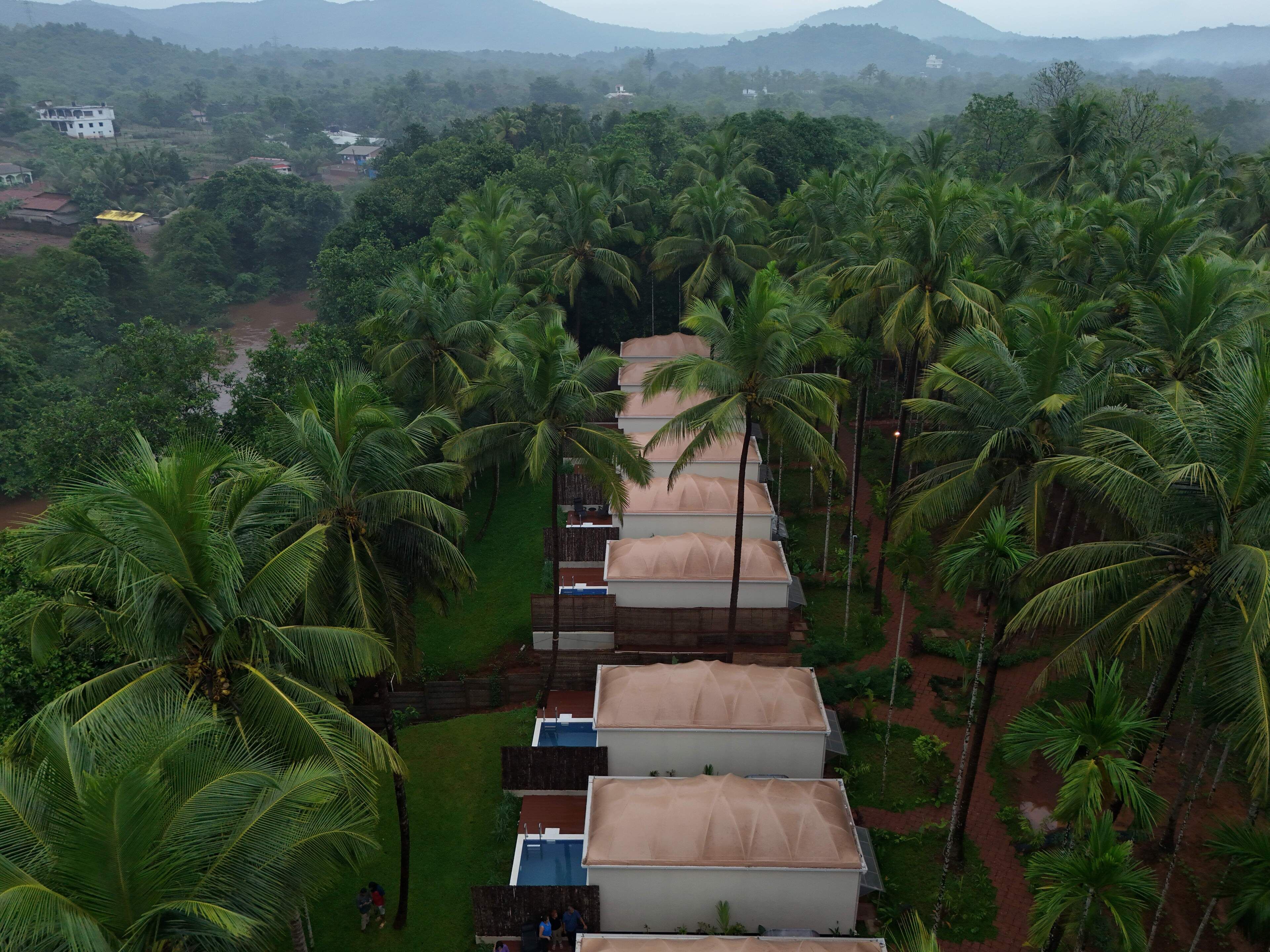 Une rangée de bâtiments à un seul étage, dotés d’une architecture de toit uniforme, est entourée d’une végétation luxuriante de palmiers, ce qui suggère un cadre tropical.