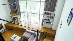 A modern living room with a sofa, a small table, and a window with a view of greenery and buildings. - Empire City Damansara M Tower by Airhost (Petaling Jaya)