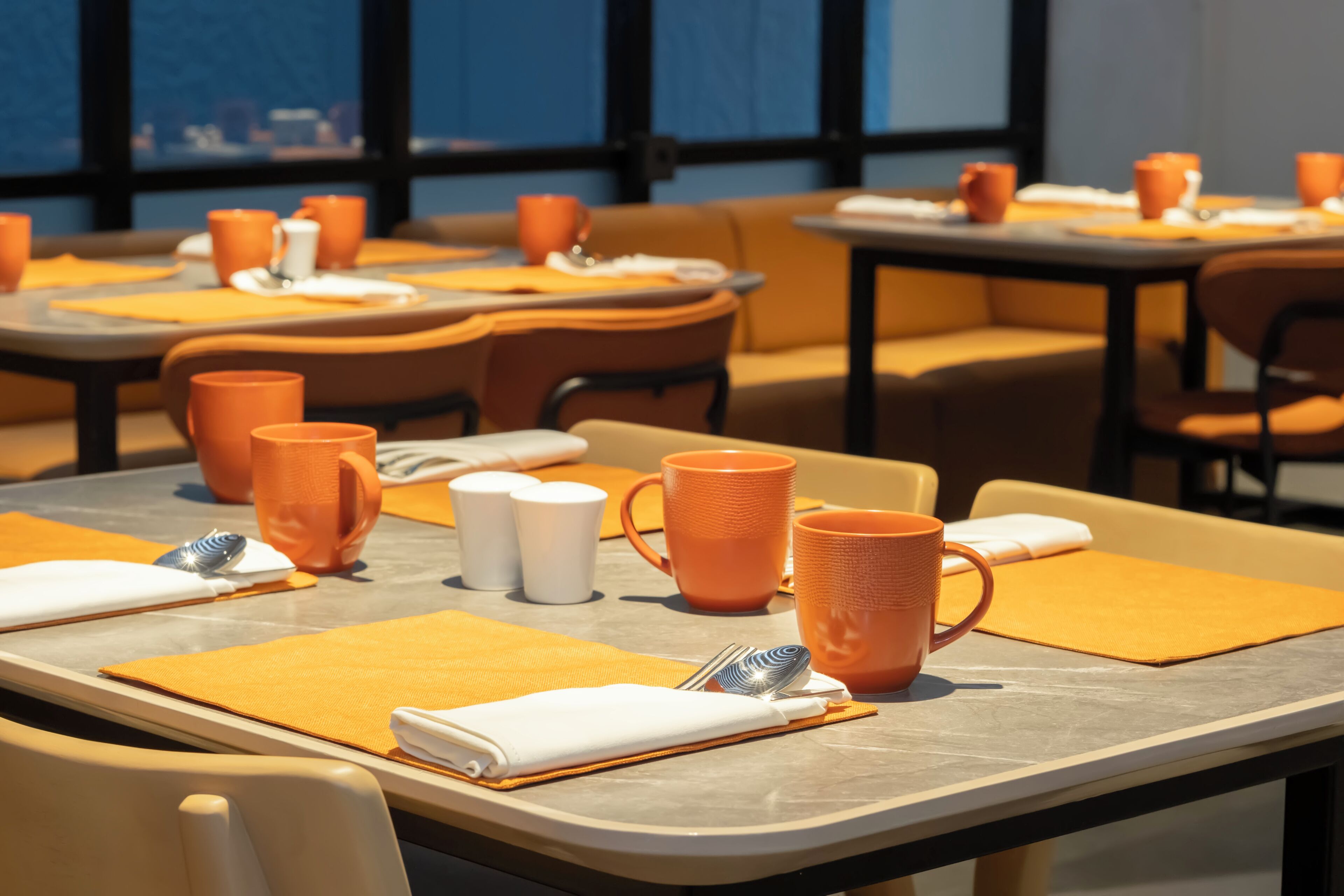 A dining area with tables set for two, featuring orange mugs, white cups, and a spoon on a yellow placemat.