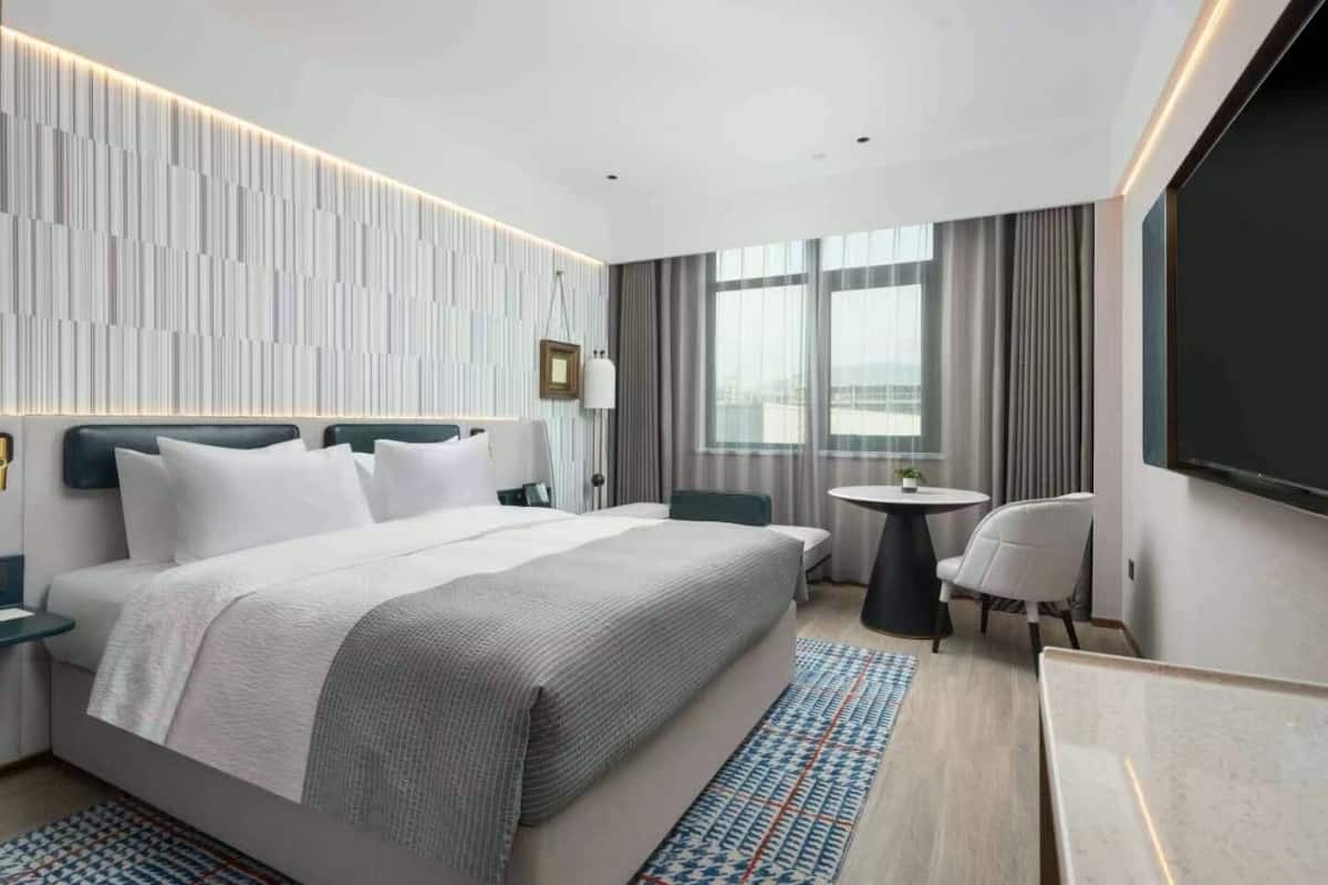 Una habitación de hotel moderna con una cama grande, una mesita redonda pequeña, una silla, un televisor y vistas de la ciudad a través de ventanas amplias.