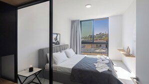 Ett modernt sovrum med en stor säng, ett sängbord med en lampa och utsikt över staden genom ett stort fönster.