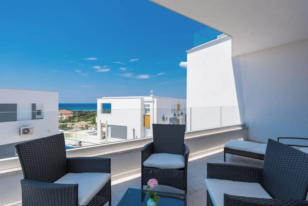 A balcony with wicker furniture, a glass table, and a view of the sea and buildings.