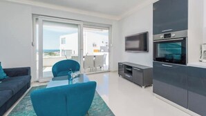 Una sala de estar moderna con un sofá, una mesa de centro de cristal y un televisor. Hay un balcón con vistas al mar.