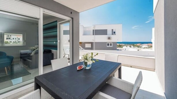 Un balcón con una mesa y sillas, un ventanal de vidrio que da a una habitación y vistas a edificios y al mar.