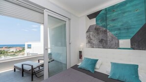 Un dormitorio moderno con una cama grande, dos almohadas, un ventanal de vidrio que da a un balcón y vista al mar.