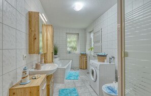 Ein modernes Badezimmer mit einem Holzkosmetikschrank, einem Waschbecken, einer Badewanne, einer Waschmaschine und einem Fenster mit Blick auf Grünflächen.