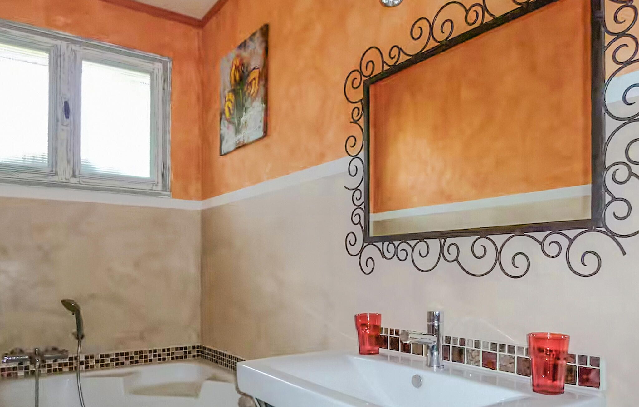 A bathroom with a bathtub, a sink with red glasses, and a decorative mirror with a black frame.